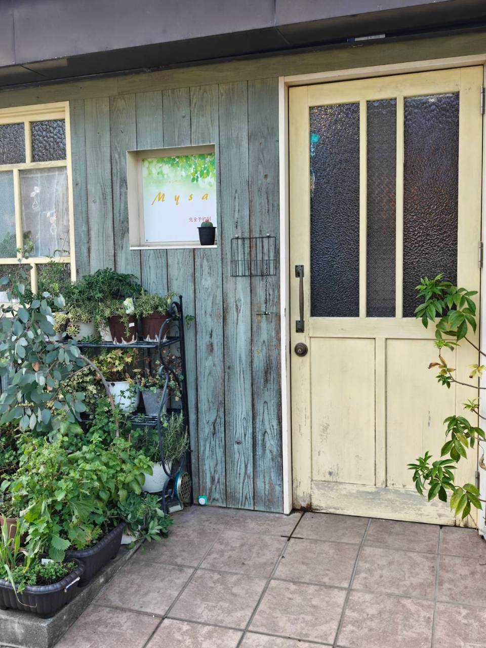 mysaの入口。水色の板壁に「Mysa」の看板、クリーム色のドアと鉢植えの植物